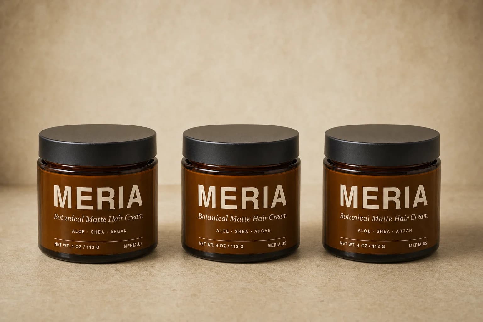 Three jars of Meria Botanical Matte Hair Cream standing in a row on a warm tan surface.